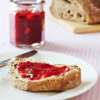 Verse aardbeienconfituur op een snede brood, met confituurpot op de achtergrond