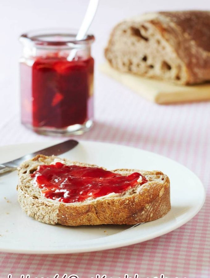 Verse aardbeienconfituur op een snede brood, met confituurpot op de achtergrond