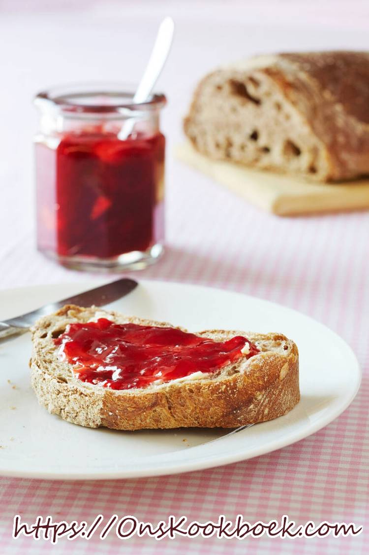 Verse aardbeienconfituur op een snede brood, met confituurpot op de achtergrond