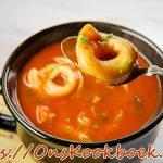 Close-up van tomaten tortellini-soep in een zwart kommetje, met een lepel die één tortellini optilt en verse groene kruiden op de glanzende tomatenbasis.