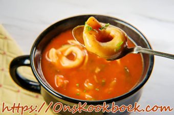 Close-up van tomaten tortellini-soep in een zwart kommetje, met een lepel die één tortellini optilt en verse groene kruiden op de glanzende tomatenbasis.