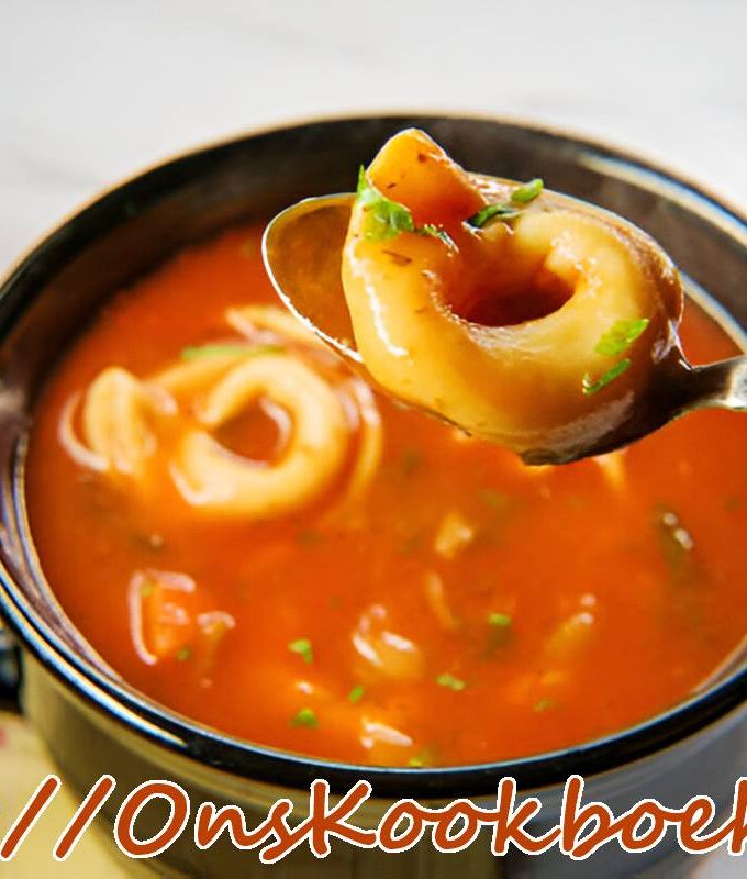 Close-up van tomaten tortellini-soep in een zwart kommetje, met een lepel die één tortellini optilt en verse groene kruiden op de glanzende tomatenbasis.