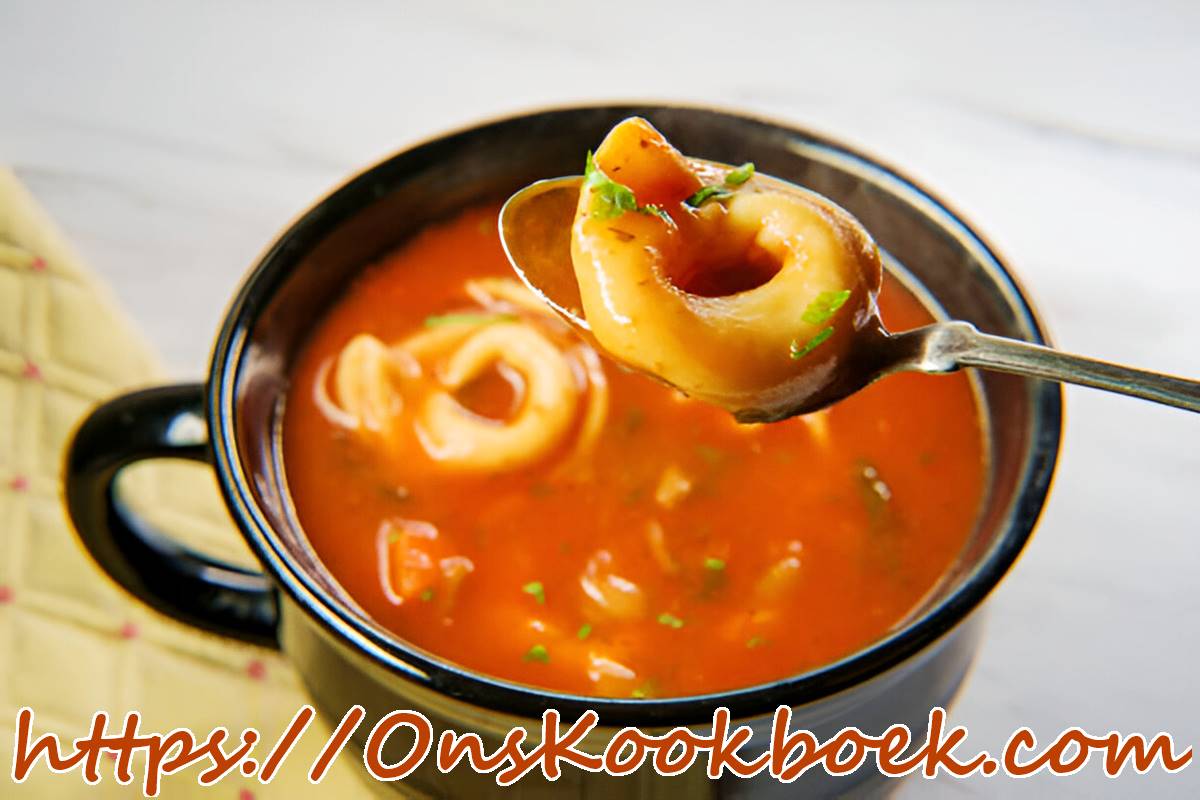 Close-up van tomaten tortellini-soep in een zwart kommetje, met een lepel die één tortellini optilt en verse groene kruiden op de glanzende tomatenbasis.